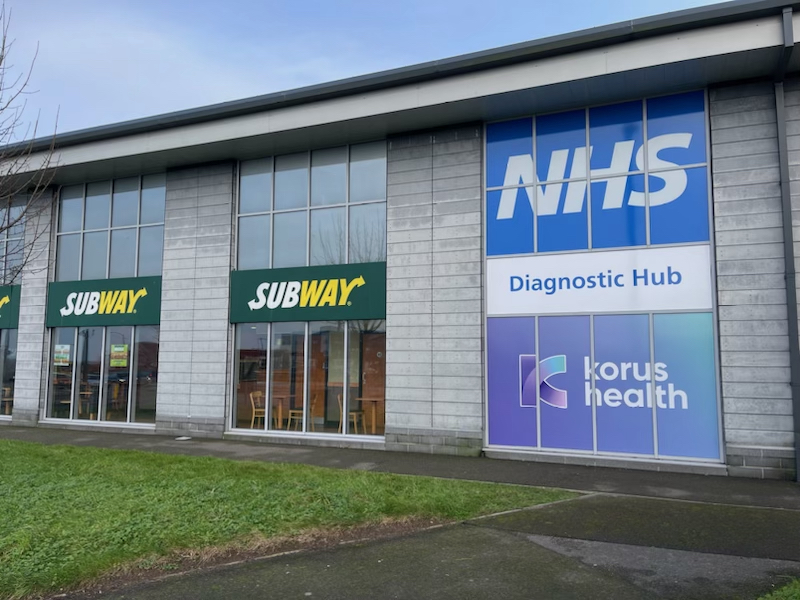 a photo of the outside of the Pool Diagnostic Hub, next door to a Subway sandwich store. The building is industrial looking with grey tiles, and large glass windows. The windows are covered in signage, saying NHS, Diagnostic hub and Korus Health